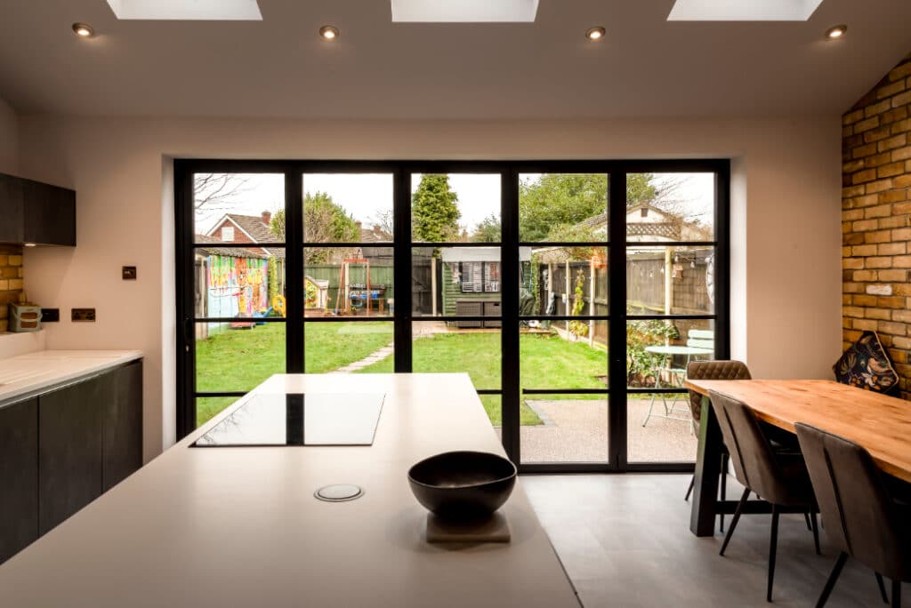 Modern kitchen with a white island, black bowl, and dining table. Large glass doors open to a garden with a mural, lawn, and furniture. Brick wall adds texture.