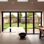 Thumbnail of http://Modern%20kitchen%20with%20a%20white%20island,%20black%20bowl,%20and%20dining%20table.%20Large%20glass%20doors%20open%20to%20a%20garden%20with%20a%20mural,%20lawn,%20and%20furniture.%20Brick%20wall%20adds%20texture.