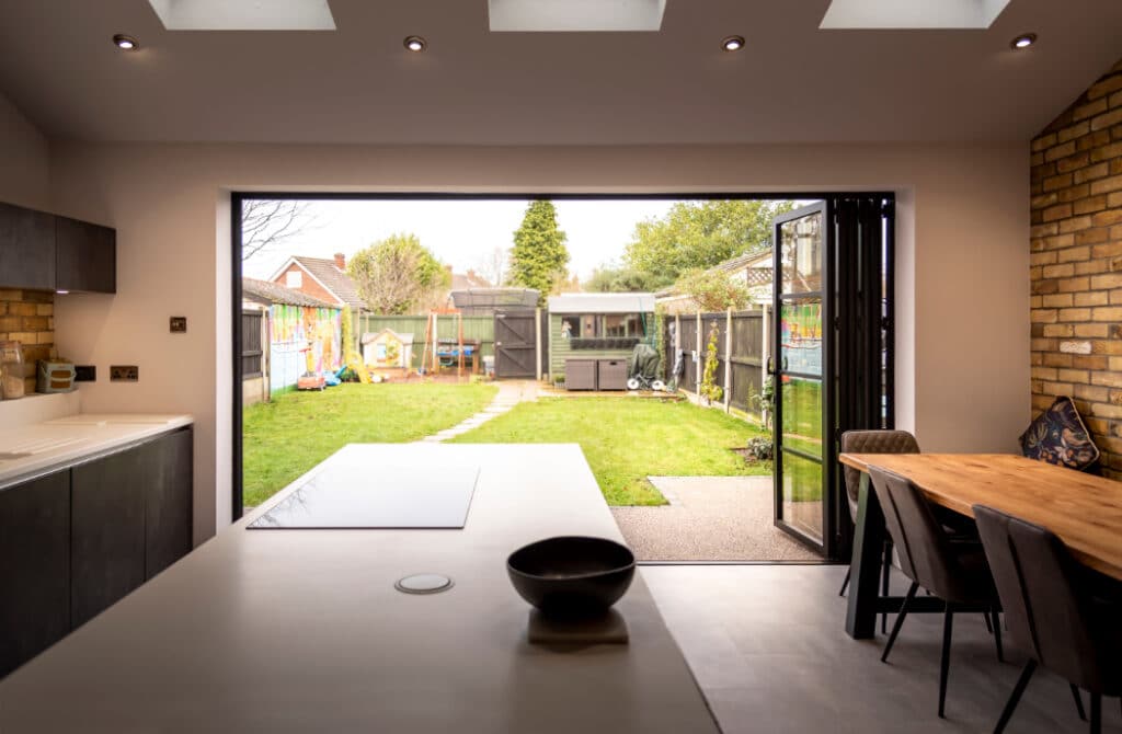 Modern kitchen with island and adjacent dining area. Bi-fold doors open to a fenced backyard with a small path leading to a shed and garden. Skylights illuminate the space.