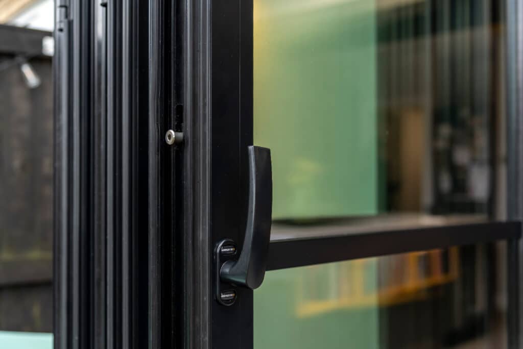 Close-up of a partially open glass door with a black metal frame and handle, showing a blurred interior background.