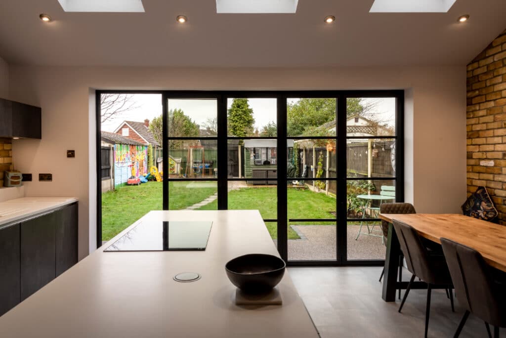 Modern kitchen with a white island and black induction cooktop. Brick wall with a wooden table and chairs. Glass doors open to a backyard with a path, shed, and colorful mural.
