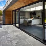 Thumbnail of http://Modern%20kitchen%20with%20glass%20sliding%20doors%20opening%20to%20a%20stone-tiled%20patio.%20Indoor%20area%20features%20a%20breakfast%20bar,%20black%20chairs,%20and%20neutral%20cabinetry.%20Outdoor%20area%20includes%20steps%20and%20green%20lawn.