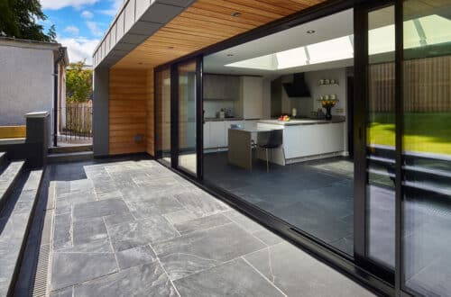 Modern kitchen with glass sliding doors opening to a stone-tiled patio. Indoor area features a breakfast bar, black chairs, and neutral cabinetry. Outdoor area includes steps and green lawn.