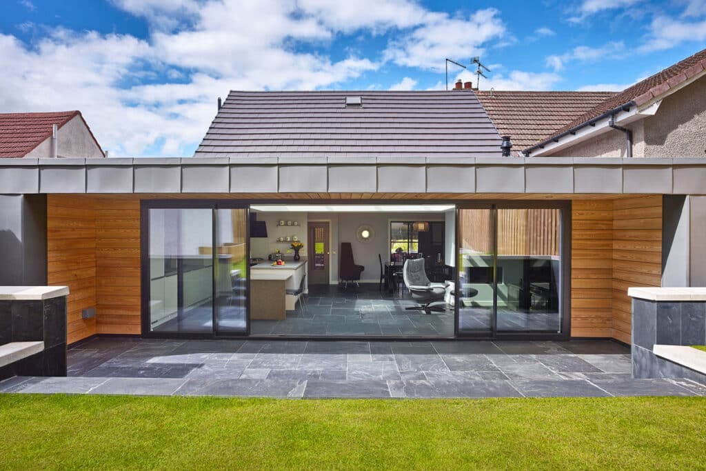Modern house with a gray tiled patio and large glass doors. The interior visible through the doors includes a kitchen and living area. The exterior walls feature wood paneling and stone accents.