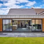 Thumbnail of http://Modern%20house%20with%20a%20gray%20tiled%20patio%20and%20large%20glass%20doors.%20The%20interior%20visible%20through%20the%20doors%20includes%20a%20kitchen%20and%20living%20area.%20The%20exterior%20walls%20feature%20wood%20paneling%20and%20stone%20accents.