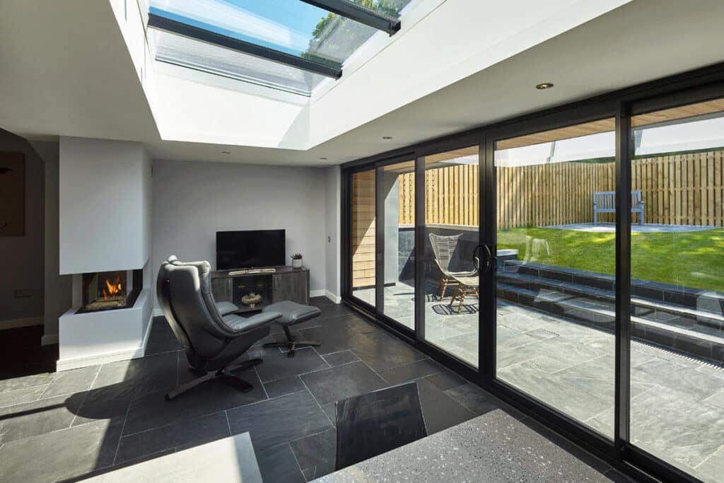 Modern living room with large glass doors leading to a patio. It features a black leather chair, a small TV, and a visible fireplace. Sunlight enters through skylights.