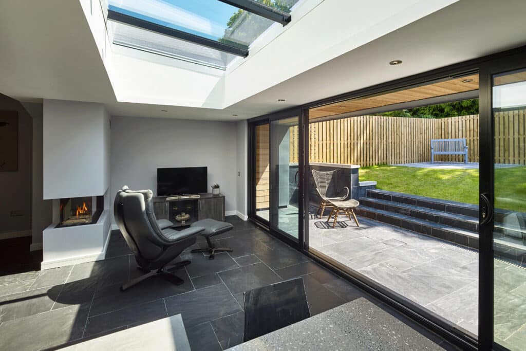 Modern room with a sleek black chair, flat-screen TV, and fireplace. Glass sliding doors open to a patio with a chair and grass area outside. Ceiling windows bring in natural light.