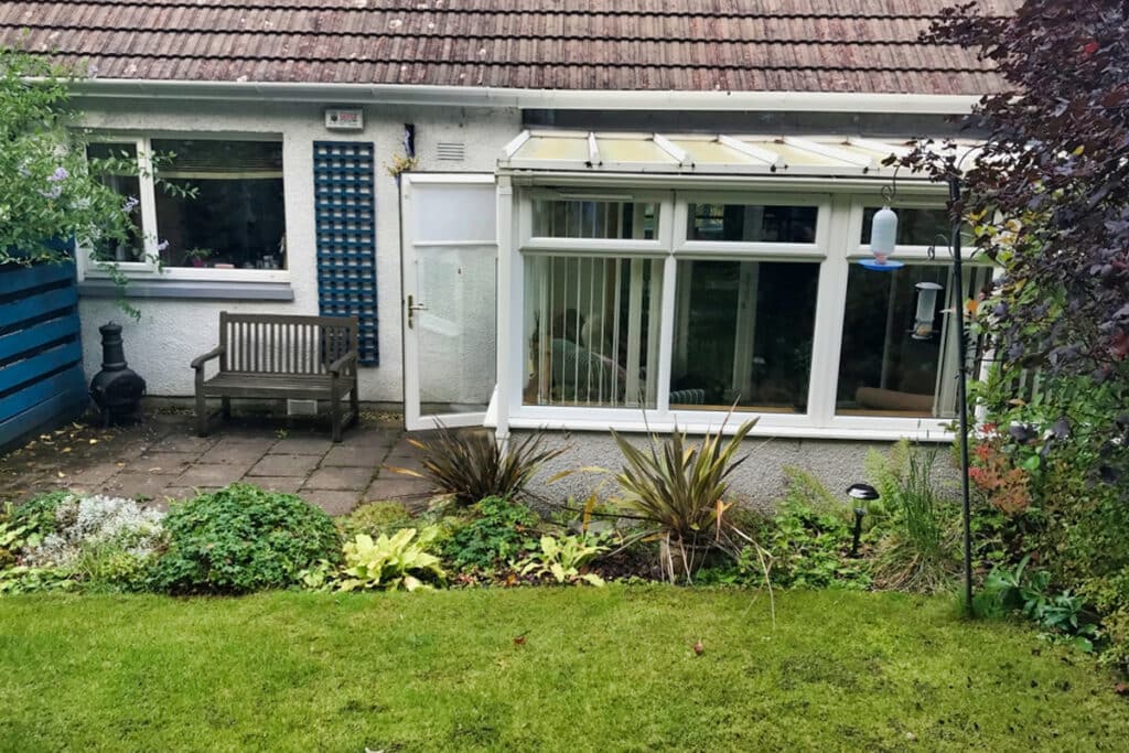 A small backyard with a patio bench, greenery, and a glass conservatory attached to a house with a tiled roof. There is a bird feeder hanging near the glass window.