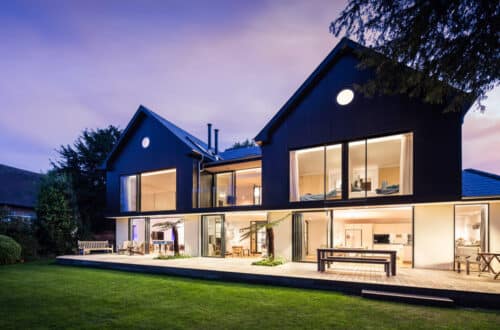 Modern two-story house with large glass windows and a wooden deck, set in a garden with a well-maintained lawn, under a twilight sky.