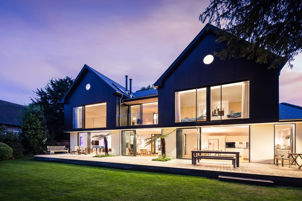 Modern two-story house with large glass windows and a wooden deck, set in a garden with a well-maintained lawn, under a twilight sky.