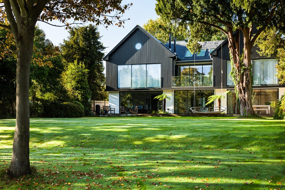 Modern two-story house with large glass windows, surrounded by trees and a well-kept lawn.