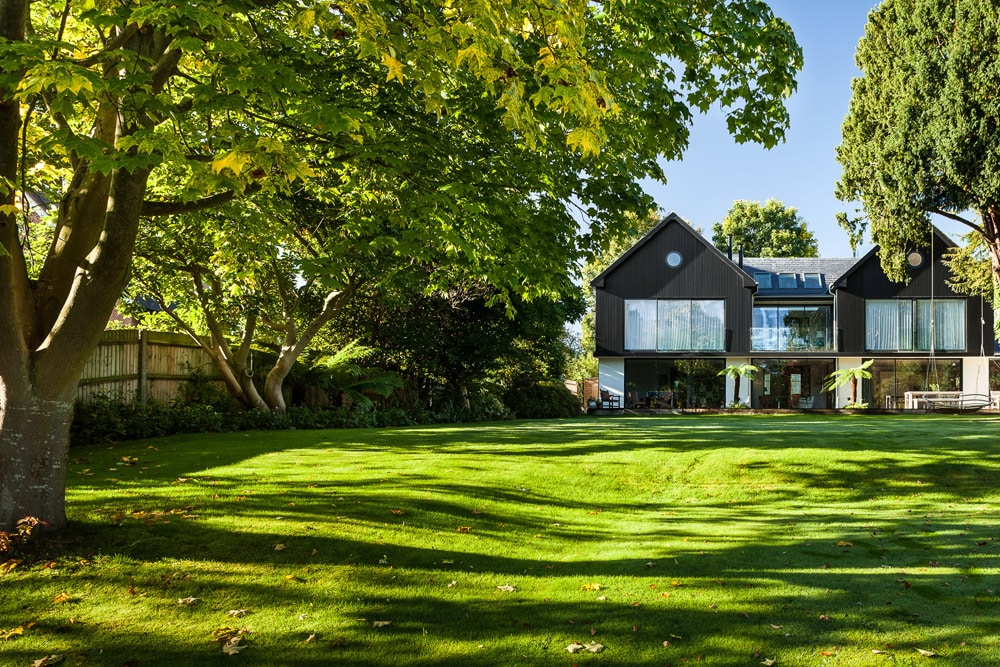 Modern two-story house with large glass windows, surrounded by lush green trees and a well-maintained lawn on a sunny day.