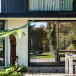 Thumbnail of http://Modern%20house%20exterior%20with%20large%20glass%20sliding%20doors,%20wooden%20deck,%20and%20a%20fern%20plant.%20Interior%20visible%20through%20glass,%20featuring%20greenery%20and%20outdoor%20reflections.