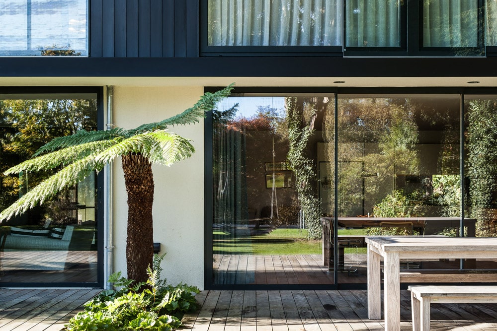 Modern house exterior with large glass sliding doors, wooden deck, and a fern plant. Interior visible through glass, featuring greenery and outdoor reflections.