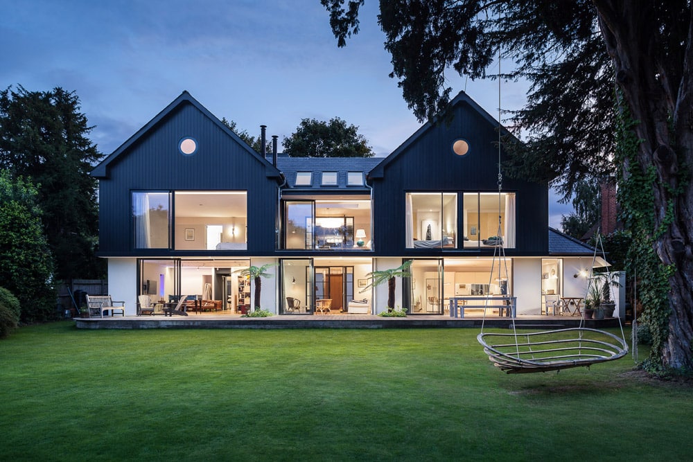 Modern two-story house with large glass windows and navy siding, featuring a spacious lawn and a hanging swing in the foreground.