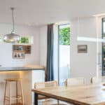 Thumbnail of http://Modern%20kitchen%20and%20dining%20area%20with%20wooden%20table,%20rattan%20chairs,%20and%20pendant%20lights%20over%20a%20black%20countertop.%20Large%20windows%20reveal%20greenery%20outside.%20Framed%20photos%20on%20the%20wall.