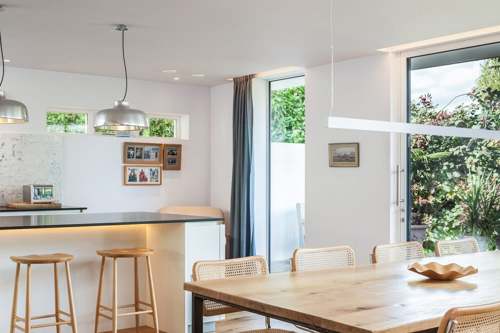 Modern kitchen and dining area with wooden table, rattan chairs, and pendant lights over a black countertop. Large windows reveal greenery outside. Framed photos on the wall.