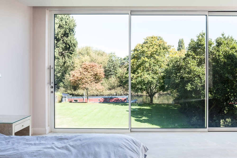 View from a bedroom with a bed and nightstand, looking through large glass doors toward a lush green lawn, trees, and a canal boat in the background.