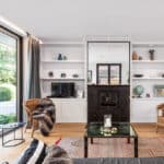 Thumbnail of http://Modern%20living%20room%20with%20glass%20doors%20opening%20to%20a%20garden.%20Features%20a%20black%20fireplace,%20wicker%20chairs,%20a%20TV,%20and%20a%20glass%20coffee%20table%20on%20a%20patterned%20rug.%20Shelves%20hold%20decorative%20items.