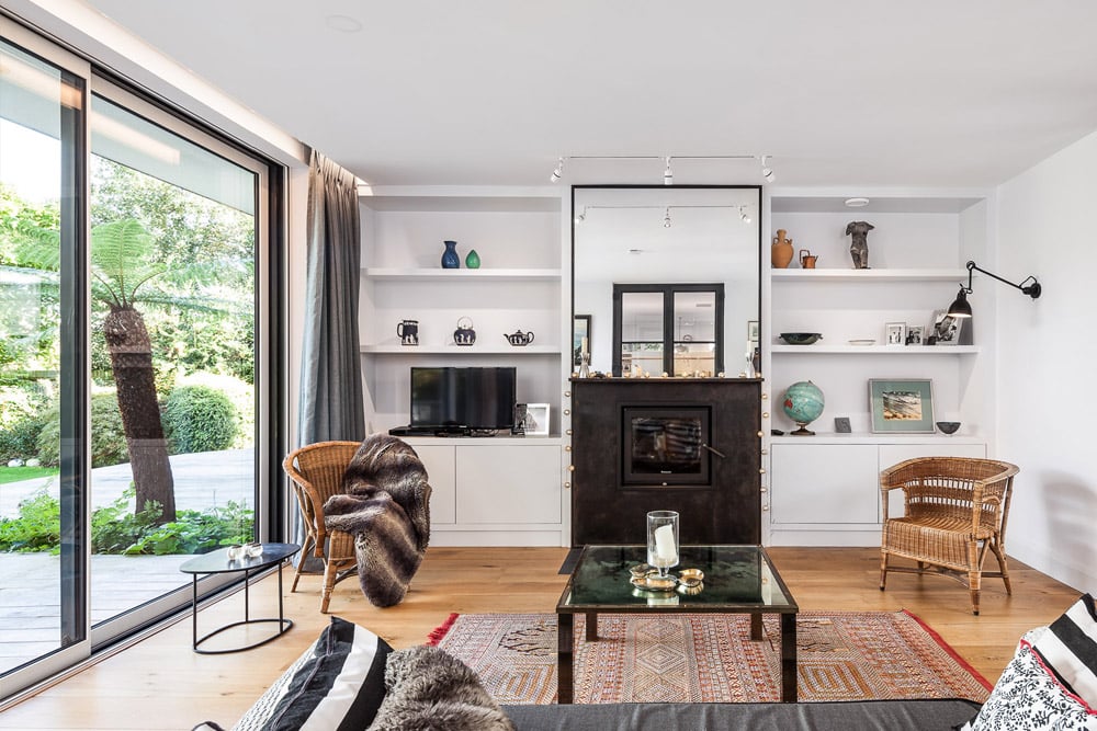 Modern living room with glass doors opening to a garden. Features a black fireplace, wicker chairs, a TV, and a glass coffee table on a patterned rug. Shelves hold decorative items.