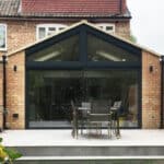 Thumbnail of http://Brick%20house%20with%20a%20modern%20glass%20extension%20and%20wooden%20slatted%20fence.%20Outdoor%20patio%20features%20a%20dining%20table%20and%20chairs%20on%20a%20concrete%20surface.
