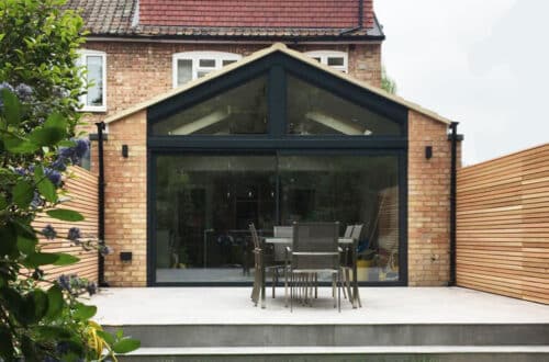 Rear view of a brick house with large windows, a gable roof, and a patio featuring a table and six chairs. A wooden fence borders the outdoor space.