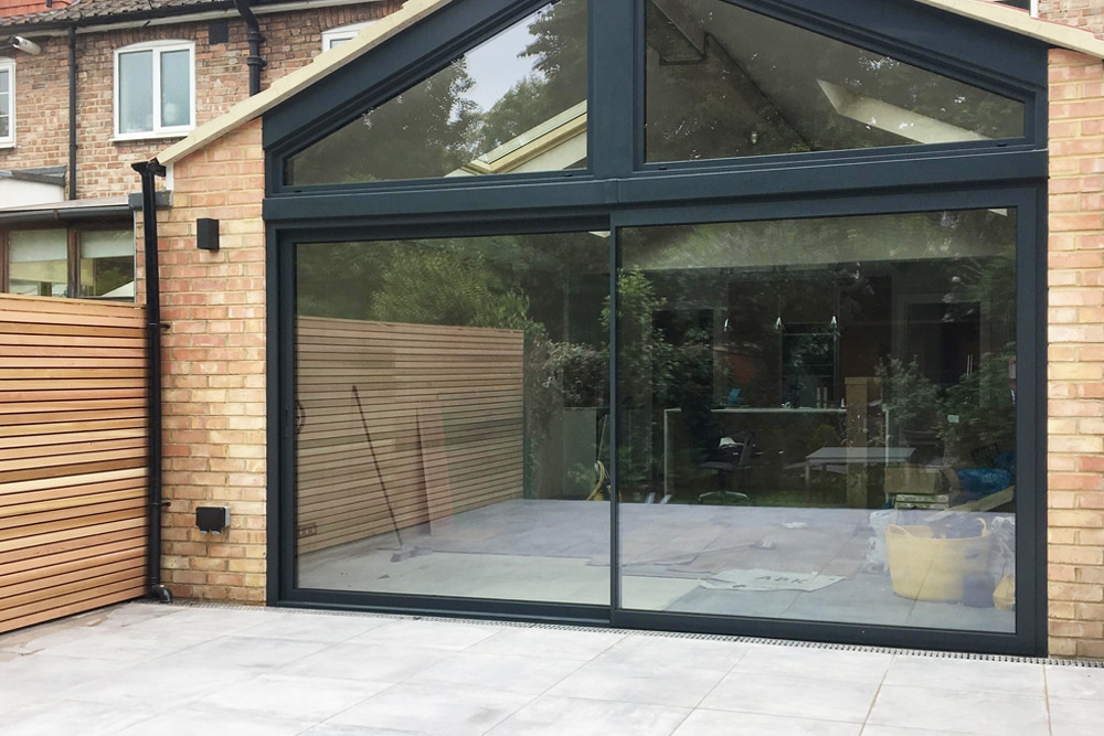 Large glass sliding doors on a brick building open to a room under construction, with tools and materials visible inside. Wood paneling adorns the outside fence.