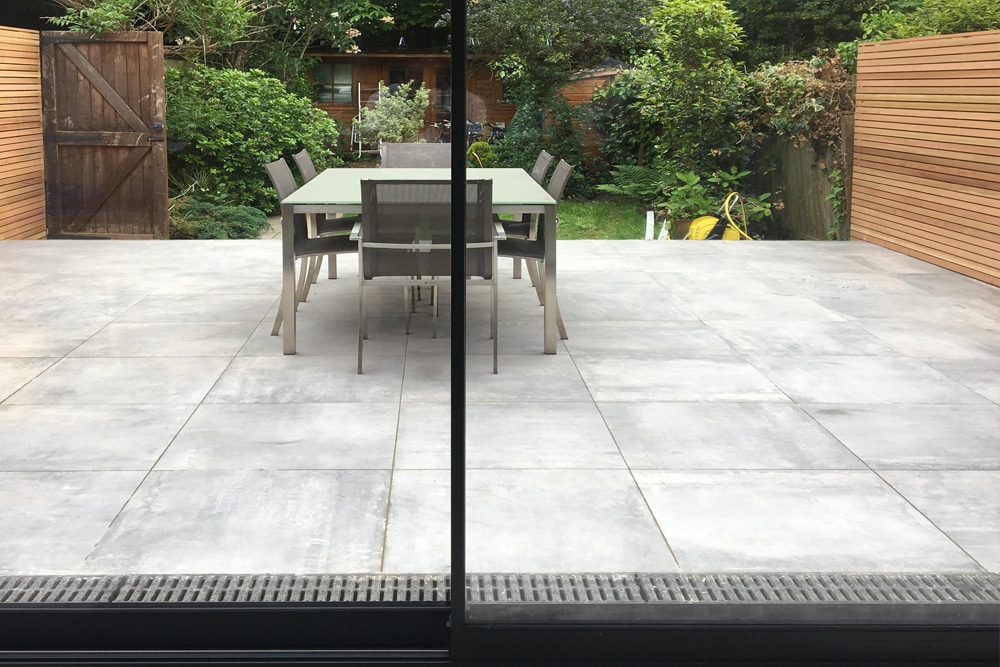 A glass door opens to a patio with a table and six chairs. The area is surrounded by greenery and wooden fencing, with garden tools visible in the background.
