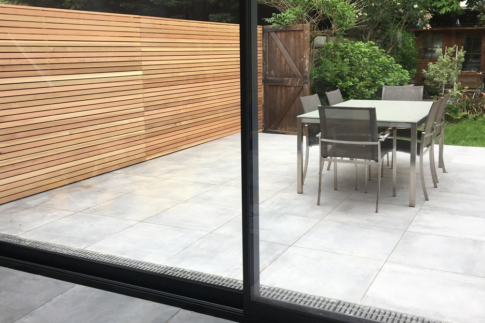 Patio with a glass table, six chairs, wooden fence, and sliding glass door. Garden with greenery and a wooden shed in the background.
