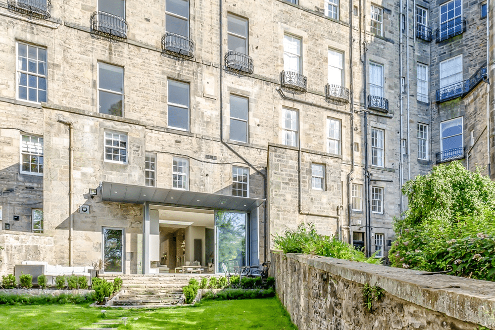 Historic stone building with a modern glass extension, overlooking a neatly manicured garden and stone pathway.