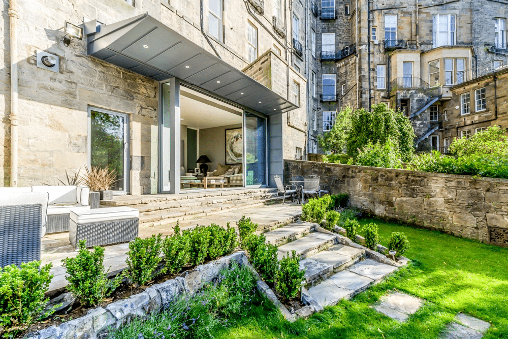 A stone building with a modern patio features sliding doors, outdoor seating, and a landscaped garden with a stone path.