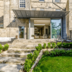 Thumbnail of http://Stone%20building%20with%20modern%20glass%20extension,%20steps%20leading%20to%20patio%20with%20outdoor%20furniture,%20surrounded%20by%20green%20lawn%20and%20shrubs.