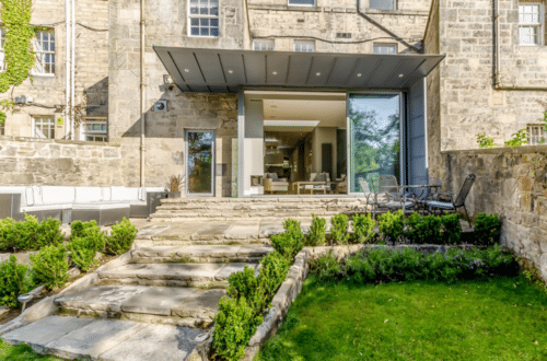 Stone building with modern glass extension, steps leading to patio with outdoor furniture, surrounded by green lawn and shrubs.