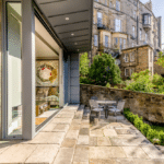 Thumbnail of http://A%20sunlit%20patio%20with%20a%20glass%20door%20opens%20to%20a%20garden,%20featuring%20a%20round%20table%20with%20chairs.%20Stone%20buildings%20and%20lush%20greenery%20are%20in%20the%20background.