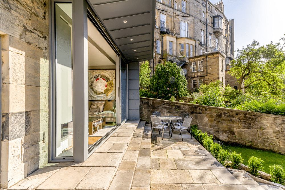A sunlit patio with a glass door opens to a garden, featuring a round table with chairs. Stone buildings and lush greenery are in the background.