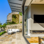 Thumbnail of http://Modern%20patio%20with%20outdoor%20seating,%20stone%20floor,%20and%20views%20of%20a%20building%20and%20greenery.%20Interior%20features%20a%20large%20TV%20and%20fireplace%20with%20glass%20wall%20opening%20to%20the%20outside.