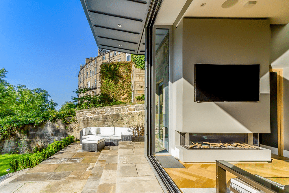 Modern patio with outdoor seating, stone floor, and views of a building and greenery. Interior features a large TV and fireplace with glass wall opening to the outside.