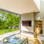 Thumbnail of http://Bright%20living%20room%20with%20large%20glass%20doors%20opening%20to%20a%20lush%20garden.%20Features%20include%20a%20wall-mounted%20TV,%20glass%20coffee%20table,%20armchair,%20and%20skylights%20casting%20natural%20light.