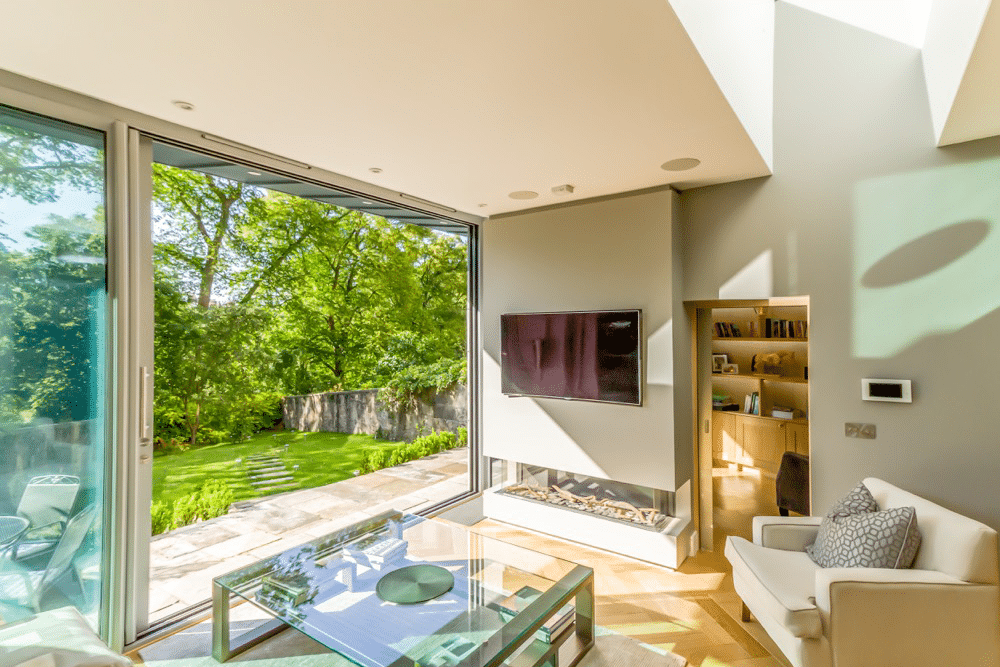 Bright living room with large glass doors opening to a lush garden. Features include a wall-mounted TV, glass coffee table, armchair, and skylights casting natural light.