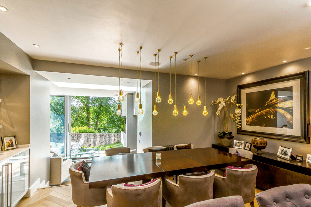 Modern dining area with a large table, hanging pendant lights, upholstered chairs, and a landscape painting. Glass doors reveal a green outdoor view.