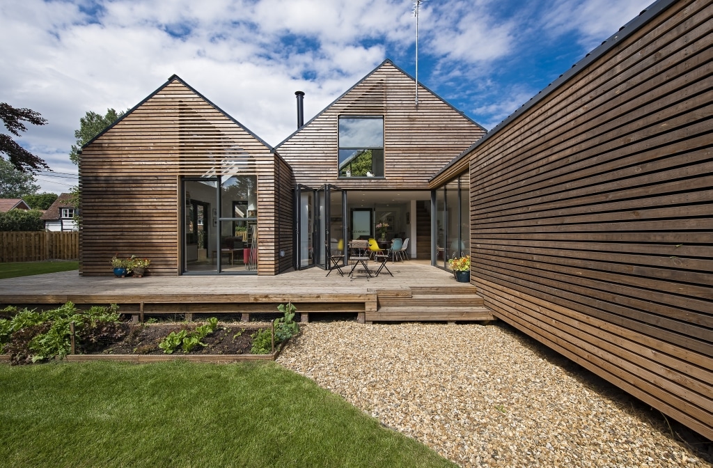 This modern house features wood paneling and large windows, complemented by a gravel path. The deck hosts an outdoor dining set, overlooking a small garden. Designed to be flood resilient, it sits gracefully under a blue sky with scattered clouds overhead.