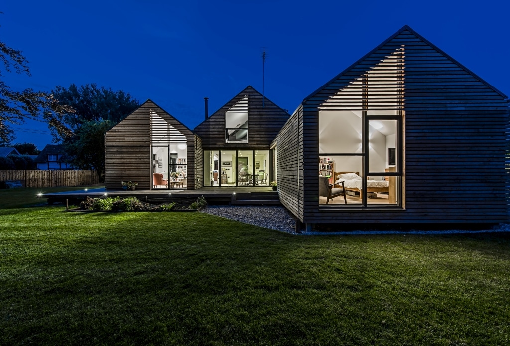 This modern, flood-resilient house boasts three sections with expansive glass windows and wooden paneling, beautifully illuminated at night. It stands gracefully amidst a dark lawn and trees, harmoniously blending form and function for sustainable living.