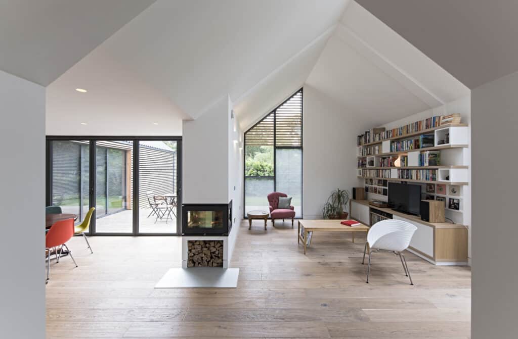 The modern living room, designed to be flood resilient, boasts large windows, a fireplace, and wooden floors. A bookshelf beside the TV complements various chairs and a red armchair in the corner. In the background, a dining area adds to its appeal.