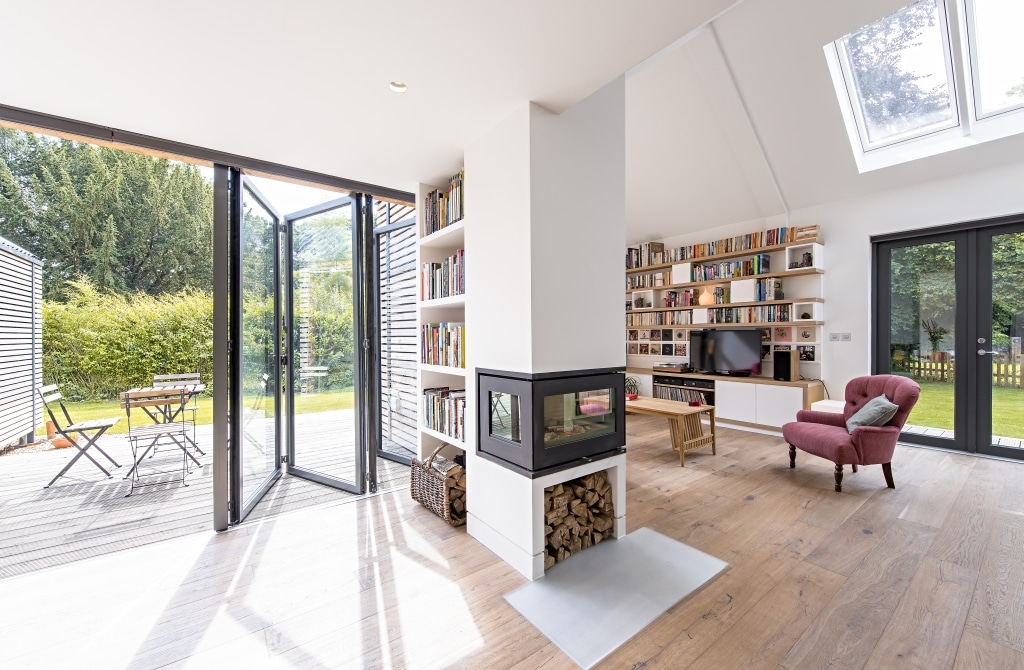 Bright modern living room with flood-resilient wooden flooring, a two-sided fireplace, bookshelves, a red armchair, and open glass doors leading to a patio with outdoor seating and a view of the garden.
