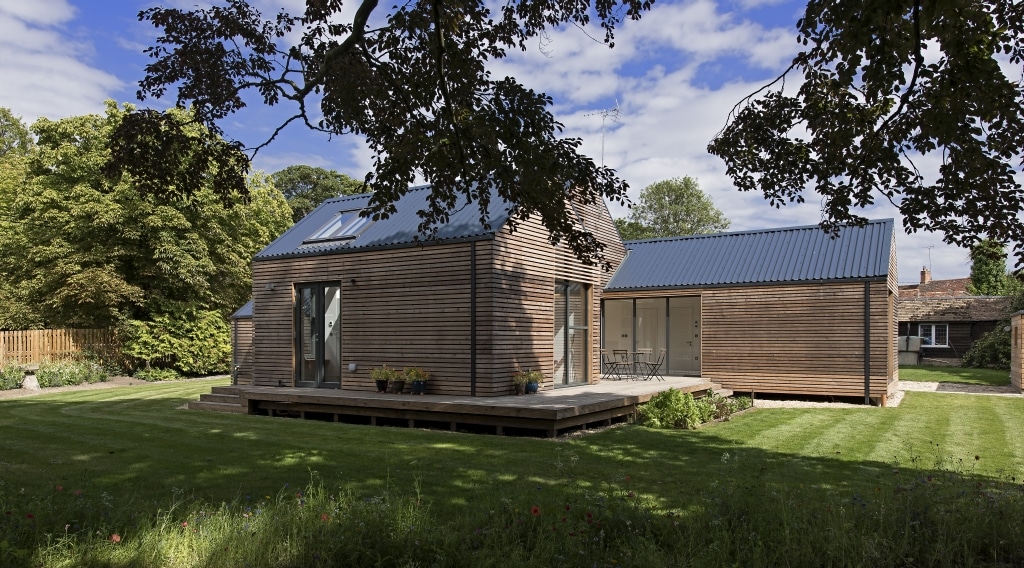 A modern, flood-resilient single-story wooden house with a blue metal roof stands proudly amid a grassy lawn and trees under a partly cloudy sky.