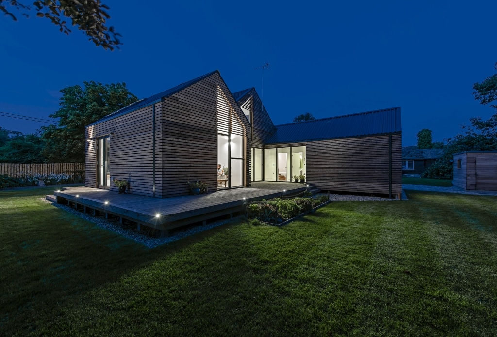 Modern wooden house with large windows, illuminated at night. Surrounded by a well-kept lawn and trees, this flood-resilient home features an angled roof and a spacious deck, effortlessly blending style with safety.