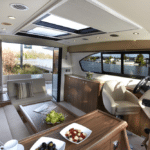 Thumbnail of http://The%20interior%20of%20a%20modern%20yacht%20features%20a%20sleek%20steering%20wheel,%20comfortable%20seating%20area,%20and%20elegant%20table%20with%20snacks.%20Large%20windows%20seamlessly%20blend%20the%20indoors%20with%20views%20of%20the%20water%20and%20trees%20outside,%20enhancing%20the%20boat's%20luxurious%20ambiance.