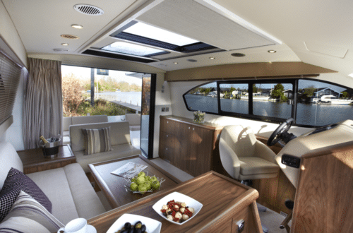 The interior of a modern yacht features a sleek steering wheel, comfortable seating area, and elegant table with snacks. Large windows seamlessly blend the indoors with views of the water and trees outside, enhancing the boat's luxurious ambiance.