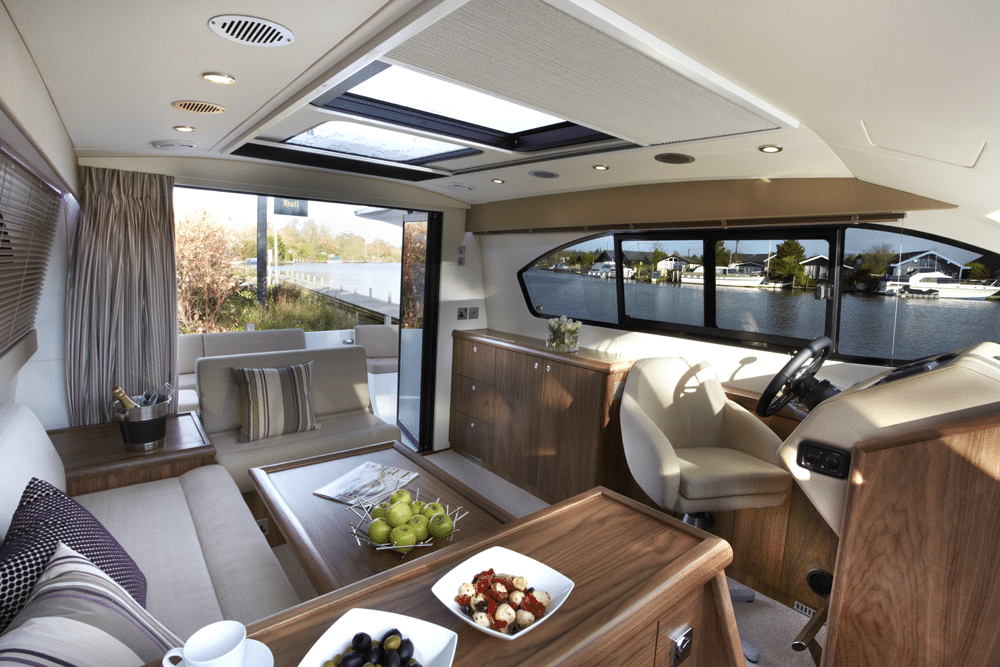 The interior of a modern yacht features a sleek steering wheel, comfortable seating area, and elegant table with snacks. Large windows seamlessly blend the indoors with views of the water and trees outside, enhancing the boat's luxurious ambiance.