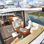 Thumbnail of http://The%20cozy%20interior%20of%20a%20docked%20boat%20features%20plush%20cushioned%20seating%20and%20a%20polished%20wooden%20table%20adorned%20with%20an%20open%20magazine.%20Wine%20and%20glasses%20await%20as%20the%20helm%20and%20controls%20stand%20ready%20inside%20the%20inviting%20cabin.